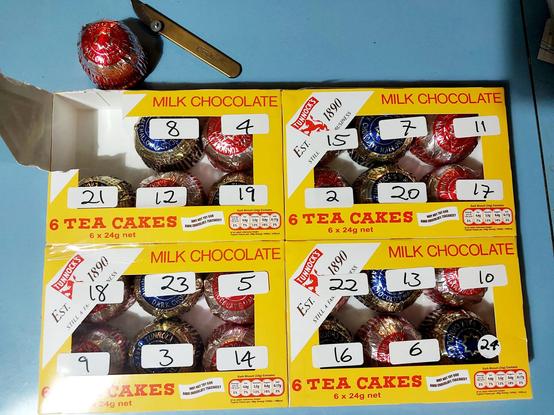 Four boxes of Tunnocks Tea Cakes, laid next to each other 2x2 on a table. Each box (which are yellow with red print, and a clear plastic viewing window) holds 6 tea cakes wrapped in foil. Handwritten labels have been stuck on the boxes above each cake, with the numbers 2-24 in a random order. There was a number 1 but that has been cut out already - a knife and a teacake are sitting on the table above the boxes.