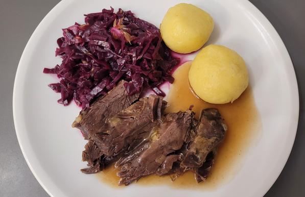 Farbfoto des beschriebenen Gerichtes: zwei gelbliche Klöße, einige Stückchen dunkles Fleisch mit relativ heller, transparenter Sauce und eine Portion Rotkohl, in der ein paar kleine Stückchen Orangenfilets zu erahnen sind. 