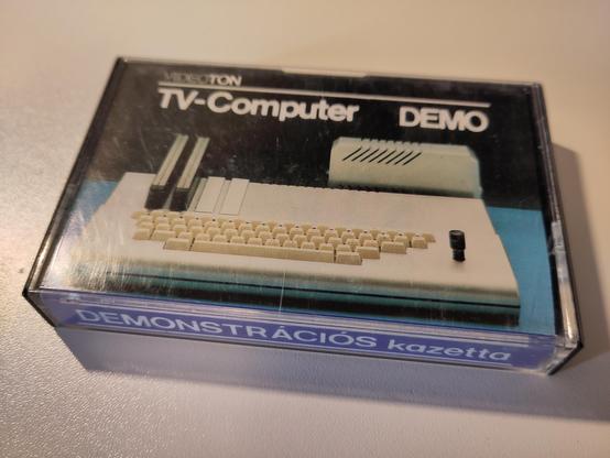 Photo of the compact cassette tape in it's case. The front side shows the marketing shot of the machine, with a "Videoton TV-Computer DEMO" label. The edge of the case says "DEMONSTRÁCIÓS kazetta".
