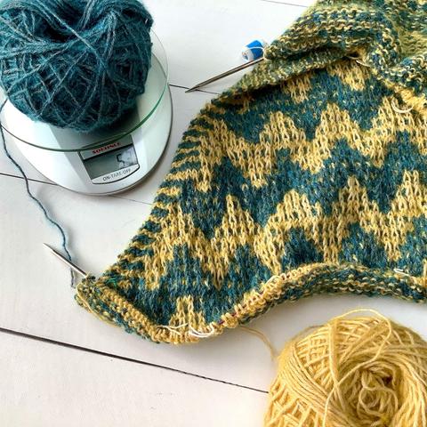 Flat lay view of a triangle shawl with mosaic patterning on a circular knitting needle, together with yellow & petrol yarn on a white plank background