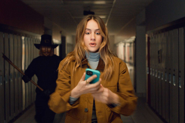 From the 2023 horror movie, Thanksgiving, we see our lead character Jessica in a high school hallway of lockers. She's facing the camera, phone in hand. Behind her right shoulder is The Carver villain, dressed as John Carver, with axe in hand.