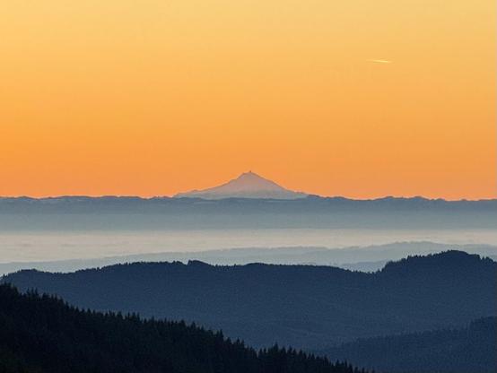 A hazy Mount Hood appears above the Cascades mountain range. The city of Portland, Oregon is in a valley of fog below. Tillamook State Forest in the foreground. Deep orange skies. Photo taken December 3, 2024. 