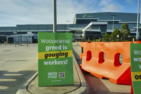 Outside Woolworths warehouse 
Union sign Text:
Woolworths’ 
GREED is 
GOUGING 
Workers
United Workers Union Photograph
IWW.org.au/woolies 