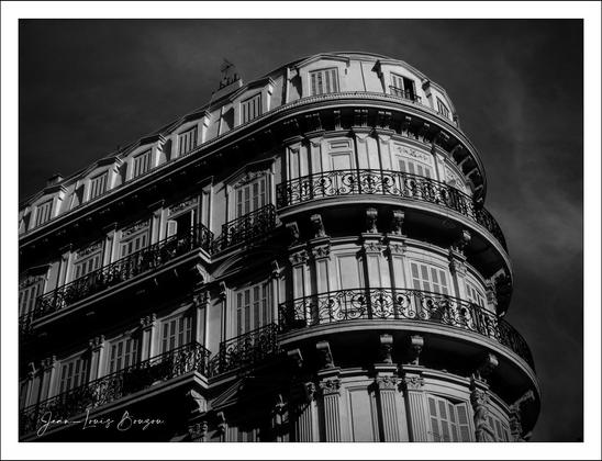 
The image depicts an elegant, multi-story building with a curved facade, captured in black and white. The architecture is ornate, featuring detailed moldings and intricate wrought iron balconies. There are multiple floors, each with tall windows that have shutters. The overall design suggests a classical or historical style, with strong shadows highlighting the building's rich textures and structural details. The sky in the background is clear, suggesting a bright day.
