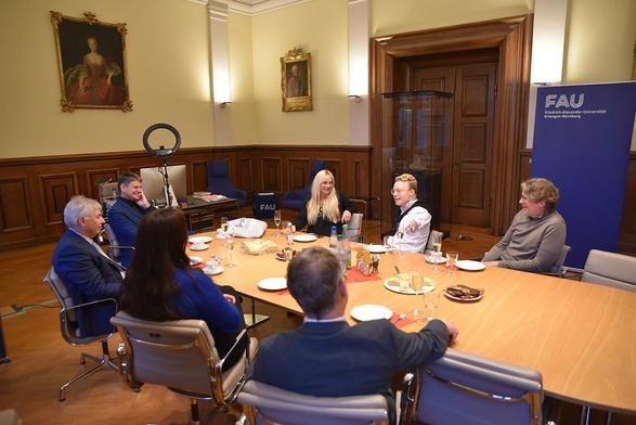Seven people sit at a conference table. Old paintings hang on the wall in the background.