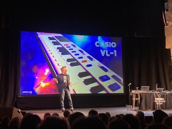 Seb on stage at the Cambridge Theatre in front of a screen with a photo of the Casio VL-1 toy keyboard on the screen behind him. 