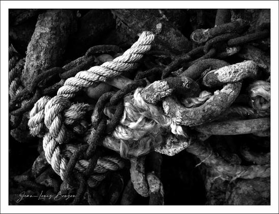 This black and white image shows a tangled mass of ropes and chains. The ropes are coiled and intertwined with rough, weathered chains, creating a textured and somewhat chaotic appearance. The overall scene appears rustic and industrial, possibly suggesting themes of strength, durability, or the passage of time.