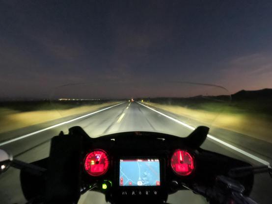 From behind the handlebars, a motorcycle rides in the late dusk grayness on a straight desert road. Motion blurs the foreground, hi-beams light the way. In the distance is darkness and a few distant lights on the horizon. The thin clouds far away glow with the last red-brown rays of late dusk against the dark, starless sky