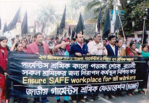 NGWF garment workers union members march with black flags while officers are holding a banner 
ENSURE SAFE WORKPLACE FOR ALL WORKERS