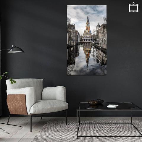 Een foto van de Waag in Alkmaar, gezien van de Witte Brug, die ligt over de Zijdam. Het water is spiegelglad zodat er een strakke spiegeling is van de waag in het water. Links en rechts zijn oude panden van de oude stad te zien. Het is verder half bewolkt, droog en windstil. 