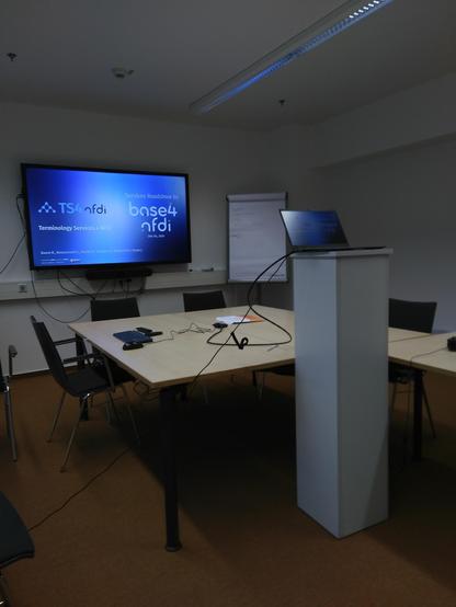 A seminar room with some tables in the middle. A huge tv screen is attach at the wall. On the screen is the introduction slide for the talk about Terminology Services 4 NFDI. In the front next to the table is an improvised speaker's desk with a notebook on it.