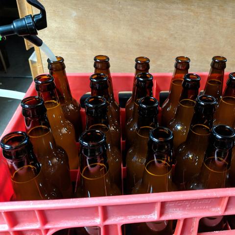 A photo of the bottles in the bottle crate, the far left bottle has the filling wand in it which is made from a picnic tap faucet and 10mm od, 6mm id silicone tubing. The bottles are filled to the top and when the wand is removed a uniform and proper amount of headspace is left. 18 bottles are visible.