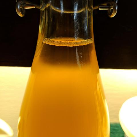 A closeup photo of beer in a clear swing top bottle. It is back lit and is a light amber color but cloudy. There are small bubbles on the surface in the bottle because it is fermenting the added dry sugar which will create carbon dioxide and pressure in the bottle.