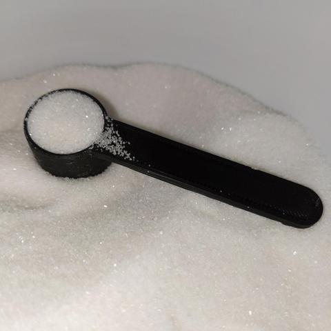 A closeup photo of a 3d printed measuring scoop that measures exactly 3.0 grams of white granulated sugar. It is sitting in white granulated sugar and is full of white granulated sugar. This scoop is for adding sugar to each 12 oz / 355 ml bottle that will be filled with beer. It was modified from a version that had the handle at the bottom of the scoop to print easier but was almost useless for scooping the sugar and getting a consistent weight. 