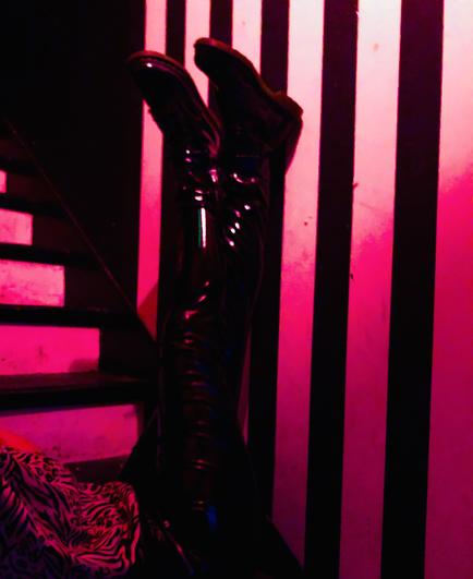 A photo taken on a staircase, a pair of legs in black pvc trousers and black patent boots are raised in the air in front of a black and white striped wall lit in red.