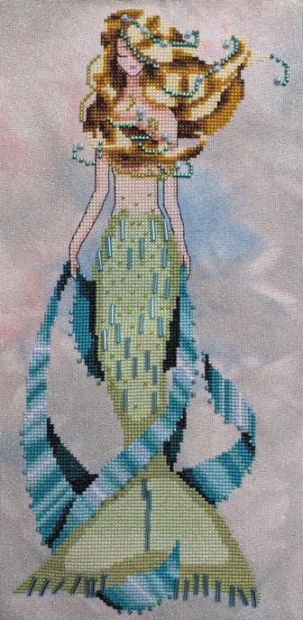 Finished cross stitch of a mermaid with a light sage green tail with a teal ribbon draped around here. There are a lot of beads to accent the pattern.