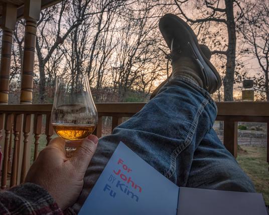 A photograph of a man's legs leading to his feet resting on a porch rail. His left hand is visible and is holding a Glencairn glass of whisky. The short story "For John" by Kim Fu is open on his lap. A small empty bottle of whisky, labeled Compass Box Delilah's XXV is next to the mans feet on the porch rail. The sky is lit with a bit of color from an almost-setting sun.