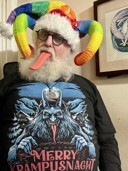 \Beardy Weirdy guy wearing a Krampus shirt with a horned, furry, red-tongued Alpine Krampus illustration with Merry Krampusnacht written on it. Also wearing a rainbow coloured Santa style hat, rainbow horns, and with a long red tongue sticking out.