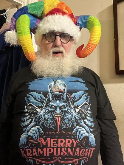 \Beardy Weirdy guy wearing a Krampus shirt with a horned, furry, red-tongued Alpine Krampus illustration with Merry Krampusnacht written on it. Also wearing a rainbow coloured Santa style hat, rainbow horns, and with a long red tongue sticking out.