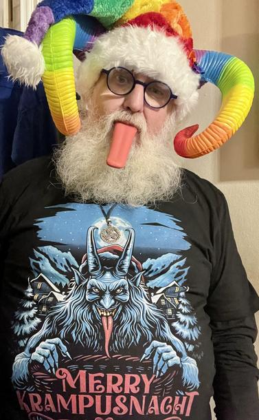 \Beardy Weirdy guy wearing a Krampus shirt with a horned, furry, red-tongued Alpine Krampus illustration with Merry Krampusnacht written on it. Also wearing a rainbow coloured Santa style hat, rainbow horns, and with a long red tongue sticking out.