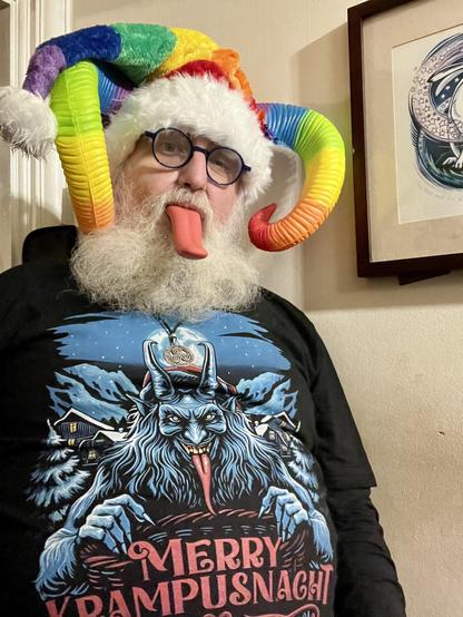 \Beardy Weirdy guy wearing a Krampus shirt with a horned, furry, red-tongued Alpine Krampus illustration with Merry Krampusnacht written on it. Also wearing a rainbow coloured Santa style hat, rainbow horns, and with a long red tongue sticking out.