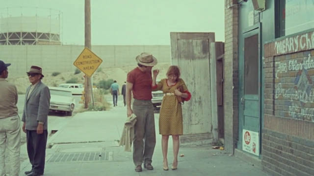 There are several people standing on the sidewalk, including a man wearing a hat and a woman in a yellow dress. They appear to be engaged in conversation. The building behind the people has a brick facade and a sign that reads "Merry Christmas" in white letters. There is also some graffiti on the wall. The image is in color, but it has a faded quality to it.