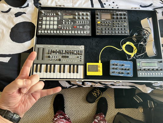 A shot from above of electronic devices arranged in a shallow rectangle on a black and white bedspread. A male left hand throws the horns because this is rocking!