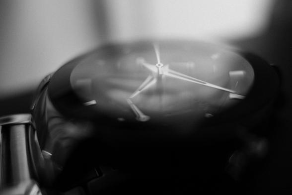 A black and white photograph of a wrist watch with a shallow depth of field.  The centre of the watch is in focus, the second hand, minute hand, and hour hand are all pointing in different directions.  The glass face of the watch is reflecting light from the room.