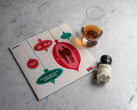 A photograph of a Glencairn glass of whisky next to a copy of a 7" record from The Both, featuring the songs "You're a Gift" and "Nothing Left to Do (Let's Make This Christmas Blue)", with a small bottle labeled "Drinks By the Dram Girvan 26 Year Old 1996 Cask 910329 The Red Cask Co." Everything is sitting on a white marble surface. 
