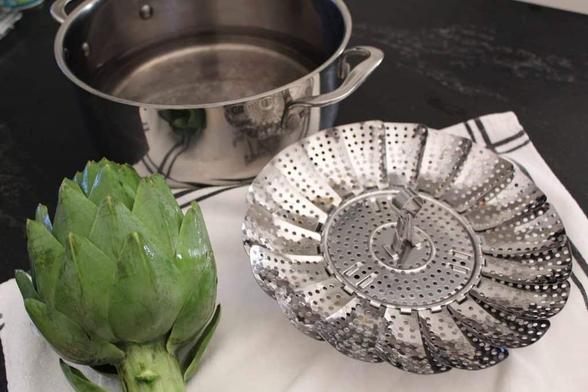 An image of a whole uncooked artichoke, a steel steaming tray with small holes that the artichoke will rest on inside a cooking pot and a steel cooking pot with a little water added for the steaming process.