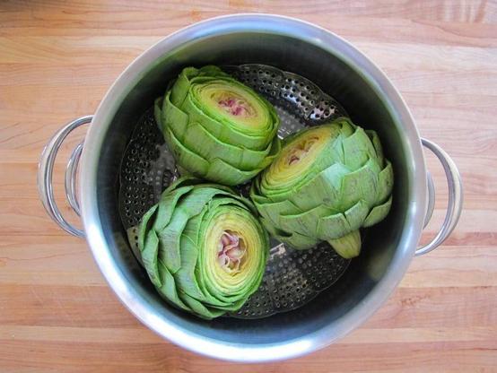 An image of 3 whole uncooked artichokes resting on their sides, atop a steaming tray and cooking pot. The top of the artichokes are sliced off, exposing their inner portions.