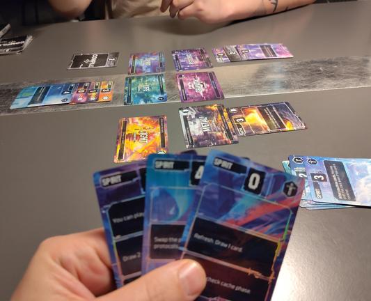 Das Bild zeigt eine Spielsituation bei einem Kartenspiel. Im Vordergrund hält eine Hand mehrere Spielkarten, die blau und farbenfroh gestaltet sind. Auf den Karten sind Texte und Symbole zu sehen, die vermutlich unterschiedliche Spielaktionen oder Werte darstellen.

Auf dem Tisch liegen weitere Karten in Reihen oder Stapeln angeordnet. Diese Karten sind ebenfalls bunt, mit verschiedenen Grafiken und Texten, die wahrscheinlich Teil des Spiels sind. Zwei Personen sitzen am Tisch und scheinen in das Spiel vertieft zu sein, wobei nur ihre Hände sichtbar sind. Der Tisch ist schlicht und grau, was den Fokus auf die Karten lenkt.