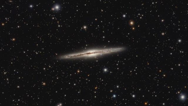 NGC 891: Silver Sliver Galaxy
Black background with stars of varying brightness and colour spread throughout.

At center running horizontally is a mostly edge-on galaxy with a slight yellow-white bulge at the middle (fills about 1/3 of the image width). Similar shape if looking at a drummers crash cymbol looking edge on.

There is also a narrow dark lane running through the middle of the galaxy (also horizontal).