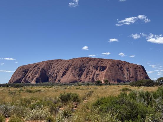 Uluru from far away during day time. Blue sky with tiny white clouds. A green desert in front.