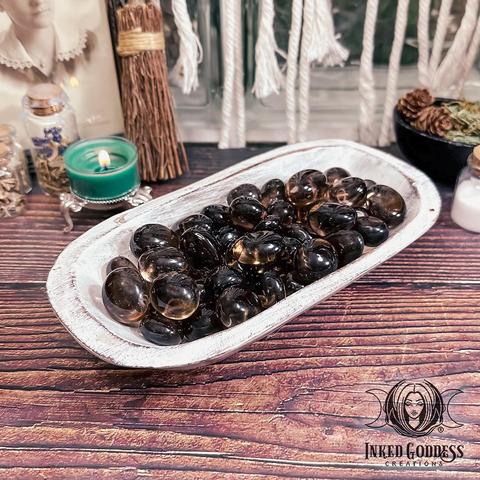 Specimens of the "Smoky Quartz AA Tumbled Gemstone" product fill a whitewashed wood bowl. They are a deep grey-brown, tumbled smooth, and are almost completely transparent. Presented by Inked Goddess Creations.