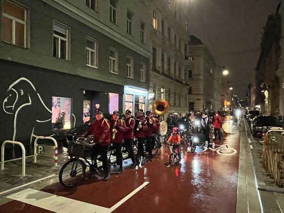 5 Musikanten des Dutch Cycling Orchestra mit Musikinstrumenten auf einem 5-Personen-Tandem, Kind in oranger Jacke auf dem Rad, dahinter viele Radfahrer-innen, rot asphaltiere Fahrbahn zur Kennzeichnung als Fahrradstraße