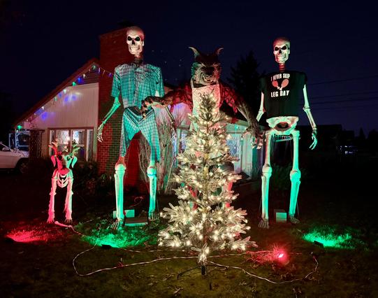 A Halloween-themed outdoor display featuring oversized skeleton figures, including one in a plaid outfit, a skeletal dog, a werewolf, with a lit Christmas Tree in front.