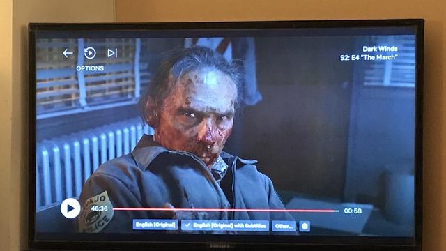 Joe Leaphorn, with bloodied facial injuries, stares intently at the camera. He is wearing a jacket with a patch indicating Navajo police. The scene is from the show "Dark Winds," Season 2, Episode 4, titled "The March”