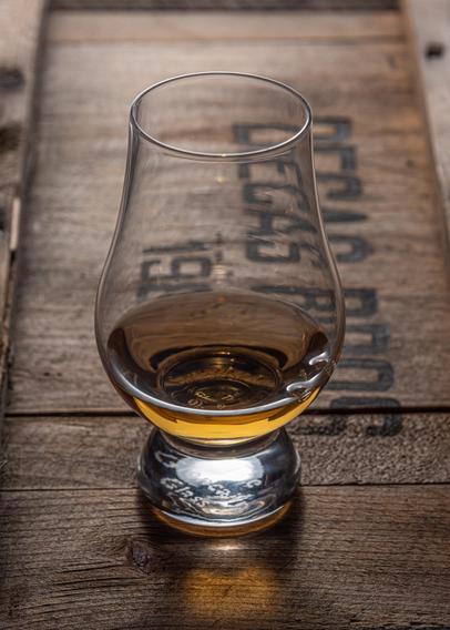 A photograph of a Glencairn glass of whisky on the face of an old cranberry crate labeled "Decas Bros 198" (with the last digit of the year obscured by the whisky.) The light is from the rear, and the whisky is casting its color onto the wood in front of the glass. 