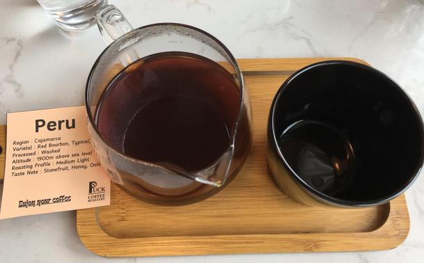 A glass jug of freshly brewed V60 coffee next to an empty cup on a wooden tray. To the left of the coffee is a piece of paper detailing the origin of the coffee: Peru, Cajamarca, Red Bourbon, washed, 1900m above sea level, Medium light, Stone fruit, honey, oolong