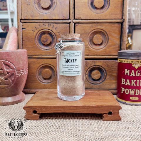 A clear glass vial of Kitchen Witch Magickal Culinary Blend: Money is displayed on a low wooden platform. The vial includes the ingredients list: organic sugar, chamomile, cloves, ginger, and nutmeg. The vial is sealed with a cork plug, and it has a green cord around its neck with a silver tree of life charm attached. This is an exclusive product from Inked Goddess Creations.