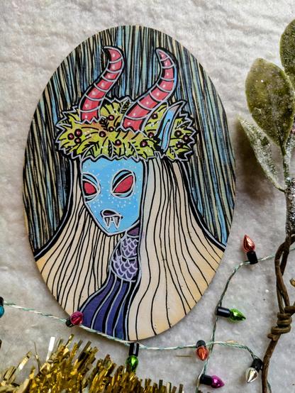 Painting on a small wooden oval of a blue-ish witch type creature with long white hair, red horns and a holly wreath.