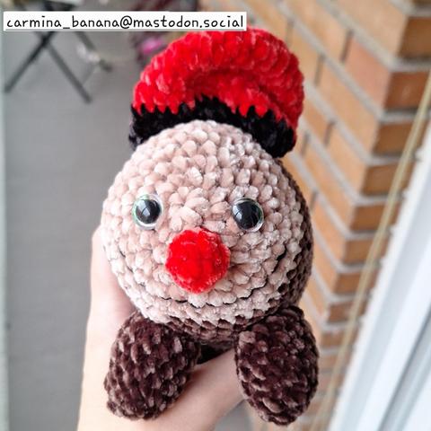 En la imagen, una mano sostiene un amigurumi o muñeco tejido a ganchillo con hilado velvet o chenilla. El muñeco representa un tió. Aquí se le ve de frente. Tiene una cara sonriente con grandes ojos verdes y una nariz roja y lleva un gorro rojo y negro