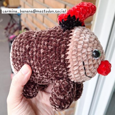 En la imagen una mano sostiene un amigurumi o muñeco tejido a ganchillo con hilado velvet o chenilla. El muñeco representa un tió. Aquí está de lado, con lo que se ven las patas delanteras y también las traseras y el perfil de la cara, junto con el gorro típico rojo y negro