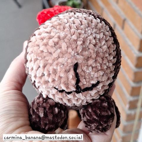 En la imagen una mano sostiene un amigurumi o muñeco tejido a ganchillo. El muñeco representa un tió. En este caso se le ve por detrás, las patas traseras y en la parte clara del tronco le han bordado un cu lo