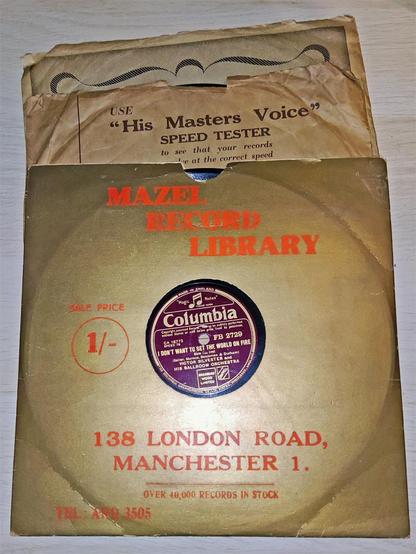 Three '78rpm' records in sleeves. The record on top has a shiny gold cover, with the words 'Mazel Record Company,  138 London Roads, Manchester 1. Over 40,000 Records In Stock. Tel: ARD 3505. Sale Price 1/- " The centre of the sleeve shows the record details, Gold lettering on a burgundy background. 