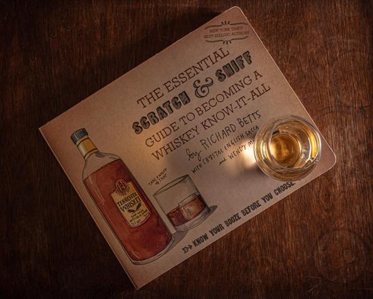 A photograph of a Glencairn glass of whisky on top of a copy of Betts, Sacca, and MacNaughton's book "The Essential Scratch & Sniff Guide to Becoming a Whiskey Know-it-All", sitting on an old dark wood surface. The light is from the right hand side and casts a shadow of the glass and whisky on the surface of the book. 