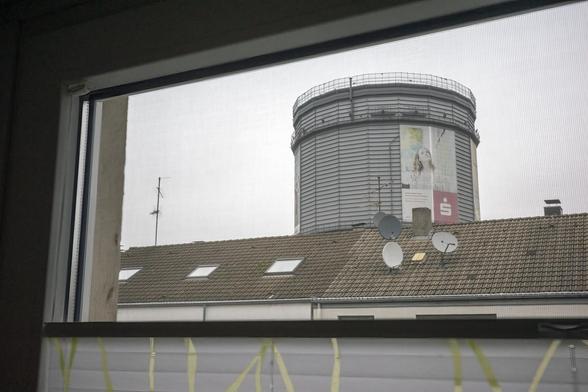 Foto: Blick durch den oberen Teil eines Fensters, teilweise verdeckt durch eine Jalousie. Zu sehen ist ein Dachfirst und ein darunterliegender Teil eines Daches, darauf Antennen und Satellitenschüsseln. Hinter dem Dach der obere Teil eines Gasometers. Es handelt sich um das Visiodrom (www.visiodrom.de) in Wuppertal-Heckinghausen.