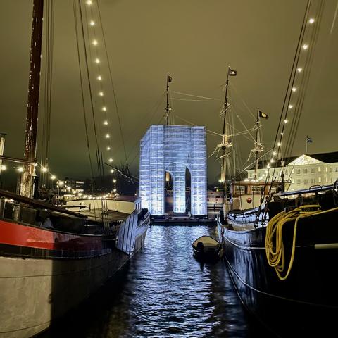 Glowing light installation on an Amsterdam canal seen through the space between two old docked ships.