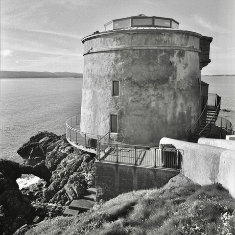 A black and white film photo. The Martello Tower Sutton stands proudly on the rocks overlooking the bay in Sutton, Dublin, Ireland. Sunday, October 22, 2023.